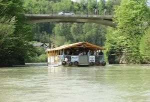 Schwimmendes Boot mit Almhütte auf einem bewaldeten Flusstal durchquert eine Brücke, auf der ein PKW und 2 Personen am Brückengelände stehen