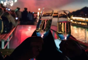 Zwei Hände mit gefüllten Sektgläsern im Abendrot mit Feuerwerk, Menschenansammlung und beleuchtetem Schiff auf Fluss im Hintergrund