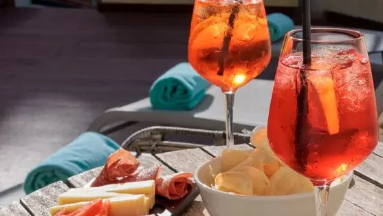 Zwei Cocktailgläser, gefüllt mit orangefarbener Flüssigkeit, je mit einem schwarzen Strohhalt und Eiswürfel, auf einem Tablett stehend, mit einer Schüssel Kartoffelchips serviert, im Hintergrund gerollte Handtücher und Sonnenliegen