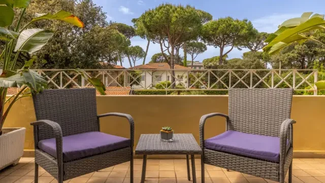 Balkonausblick auf die Balkonterrasse, mit 2 Korbstühlen mit lilafarbenen Sitzauflagen und einem Tischchen, dahinter gemauertes Balkongeländer in beiger Farbe, in Hintergrund Bäume und blauer Himmel mit weißen Wölkchen