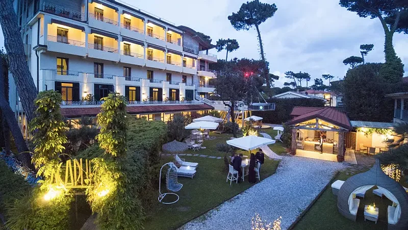 Dreistöckige Hotelanlage mit Gartenbereich, gepflasterten Wegen und Bäumen im Bildvordergrund, bei Dämmerungslicht