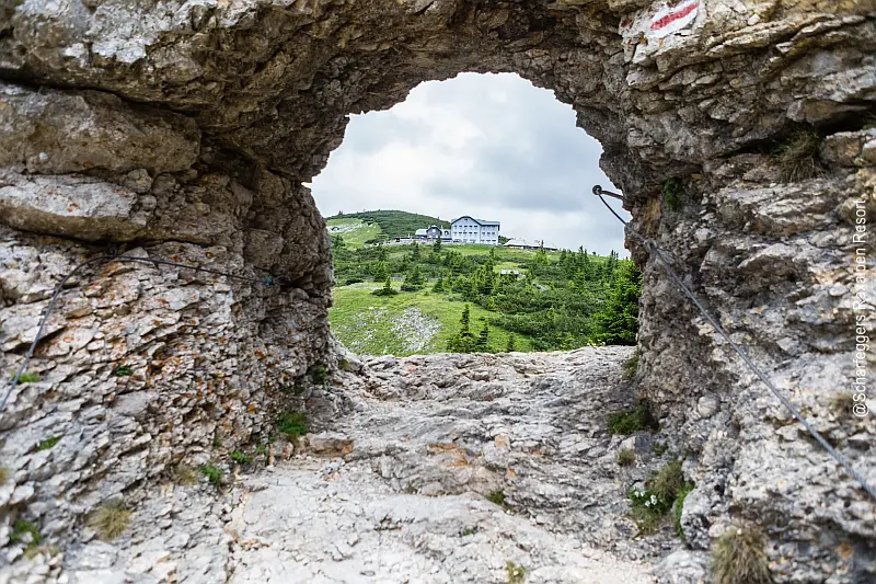 Blick aus einer Felsöffnung auf eine grüne Berglandschaft und dem Raxalm-Berggasthof in der Ferne