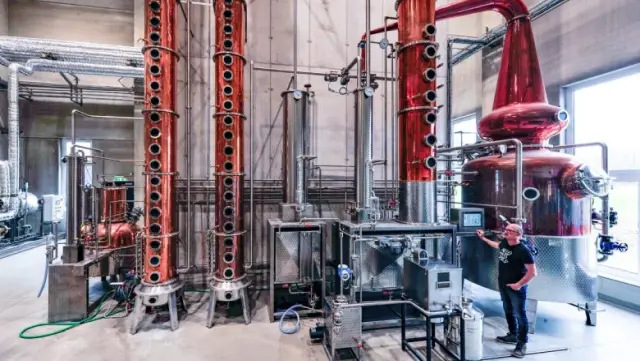 Große Destillerieanlage mit deckenhohen Kupferrohren und großen Tanks, eine Person steht im Vordergrund zum Größenvergleich