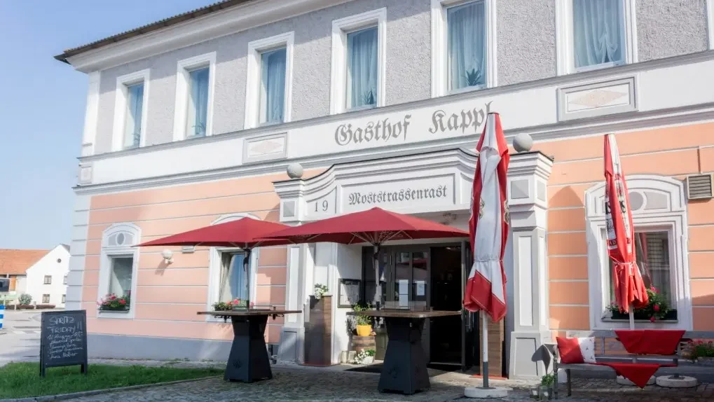 Zweistöckige Gebäudefront, Erdgeschoss rosafarben, Obergeschoss grau mit weißen Fensterrahmen, mit Aufschrift "Gasthof Kappl - Moststrassenrast", davor gepflasterter Weg, zwei Stehtische mit roten Schirmen vor dem Eingang