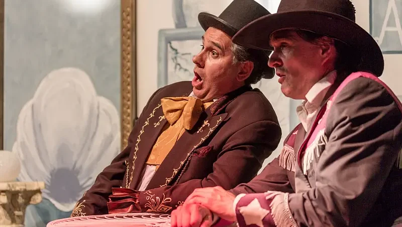 Szenenausschnitt mit 2 Schauspielern sitzend am Tisch auf einer Bühne,