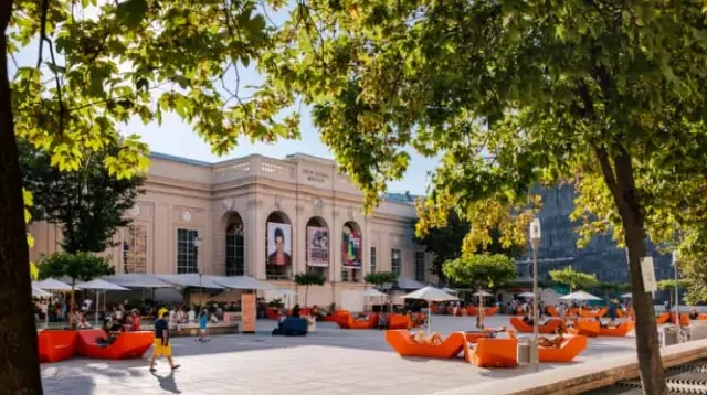 Frontansicht eines Gebäude aus der Barockzeit, bei Sonnenlicht, im Vordergrund Sitzgelegenheiten und Sonnenschirme, grün belaubte Äste eines Baumes im oberen Bildbereich
