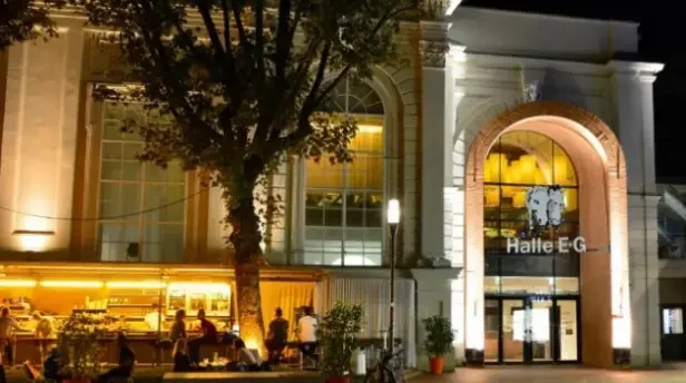 Eingang Halle E bei einem barocken Gebäude, bei Nacht, mit gelber und weißer Beleuchtung. Ein belaubter Baum mit Sitzgelegenheiten steht vor dem Gebäude