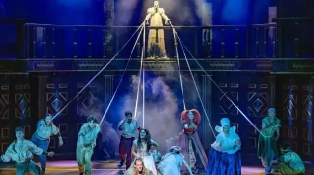 Szenenausschnitt aus dem Musical Romeo & Julia, Bühnendarstellung dex Todesengels, der über einer Gruppe von Darstellern schwebt und mit Schnüren mit diesen verbunden ist. Dunkler Hintergrund, blaue Beleuchtung.
