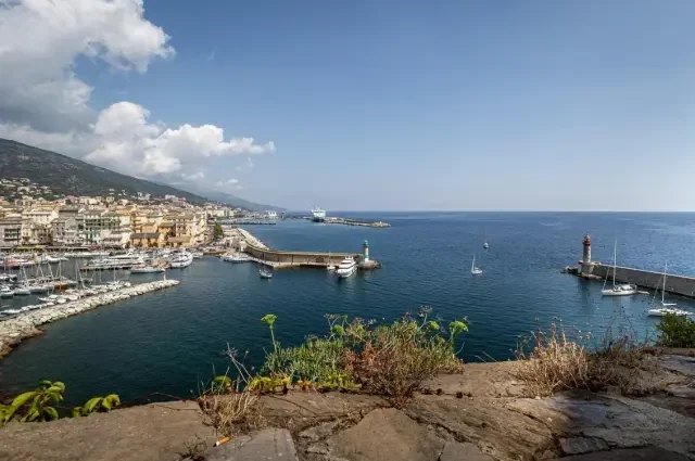 Erhöhter Blick auf den Alten Hafen von Bastia mit dicht bebauten Häuserzeilen, einer langen Kaimauer und weißen Yachten im tiefblauen Mittelmeer vor grauem Felsufer im Vordergrund.