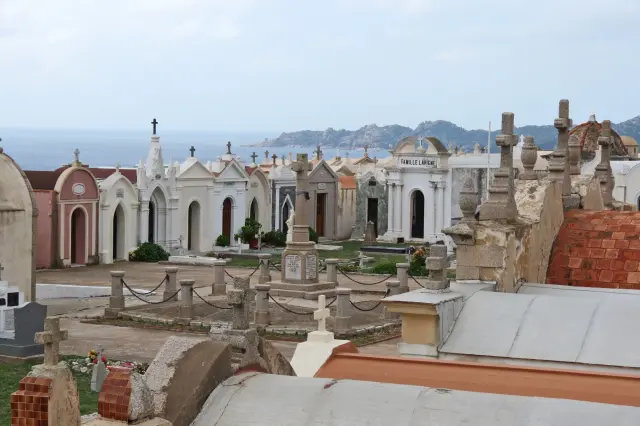 Blick über den Meeresfriedhof von Bonifacio auf Korsika mit aneinandergereihten, weißen Mausoleen und Grabkapellen im neoklassizistischen Stil vor einer hügeligen Küstenlandschaft und dem fernen Meer.