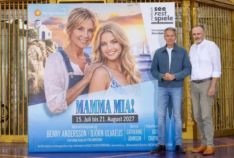 Sujetbild Musical MAMMA MIA, Plakatwand mit Foto: Porträt zweier Frauen mit blonden Haaren in Sommerkleidern, im Hintegrund Meer, Segelschiff, Insel mit typisch griechischer Kirche - weiße Fassade und blaue Tür