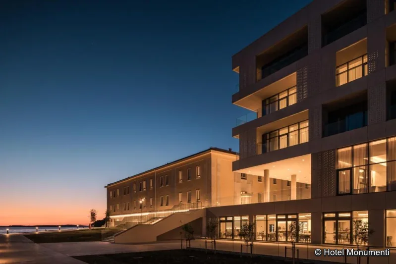 Außenansicht modernes Hotel am Meer bei Abenddämmerung, mit beleuchteten Zimmern und Blick auf die Küste.