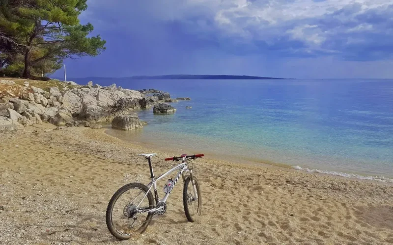 Abgestelltes Fahrrad am Kiesstrand im Bildvordergrund, Meer in verschiedenen Türkis- und Blautönen im Hintergrund, Pinie am linken Bildrand