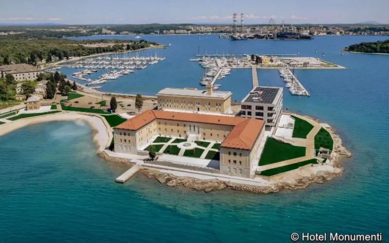 Luftaufnahme einer modernen Hafenanlage mit türkisblauem Wasser, Yachthäfen, und einem historischen Gebäudeensemble mit roten Ziegeldächern auf einer kleinen Halbinsel