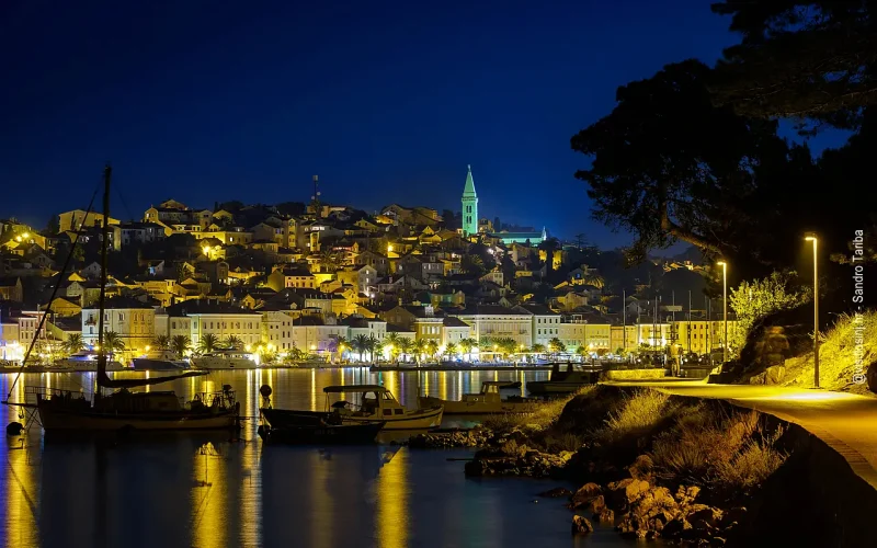 Beleuchtete Stadt bei Nacht am Meer, im Vordergrund Hafen mit kleinen Booten, im Hintergrund beleuchtete Bucht und Kirchturm