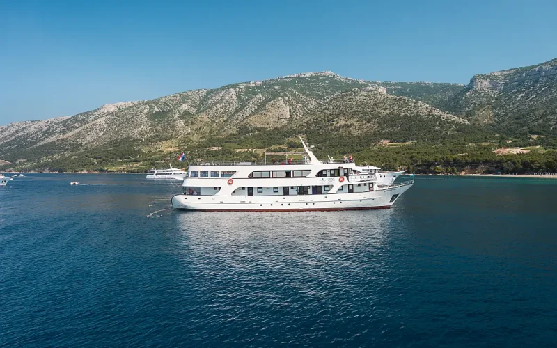 Minikreuzfahrtsschiff MS Majestic im Bildmittelpunkt spiegelt sich im Meerwasser, im Hintergrund mehrere kleine Boote und eine karge Hügellandschaft am Horizont, wolkenloses sonniges Wetter