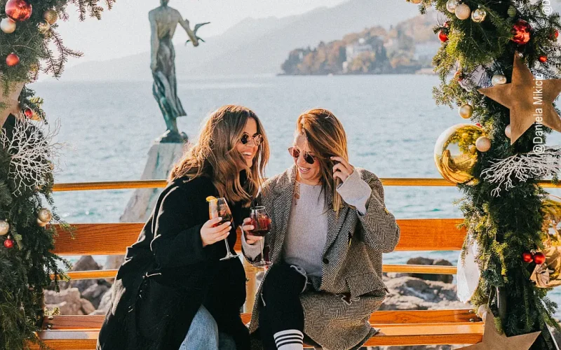 Zwei Frauen in Wintermänteln auf einer Parkbank unter einem Weihnachtsbogen an der Küste, im Hintergrund das Meer und eine Frauenstatue mit einer Möwe in der Hand
