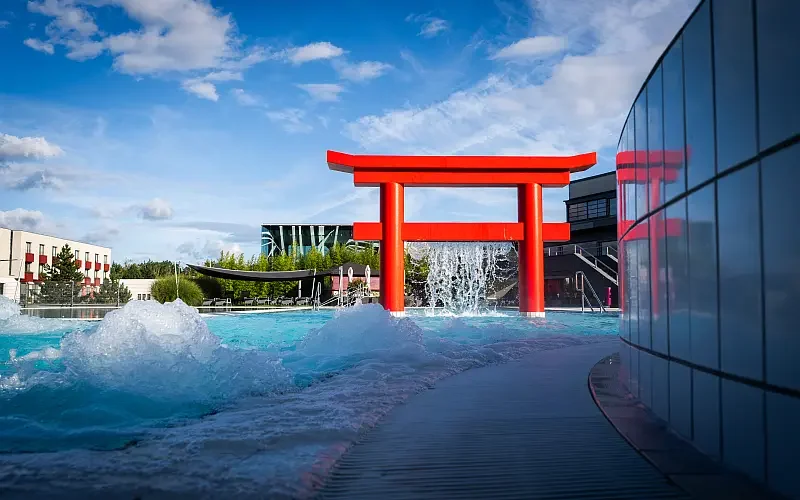 Außenpool mit sprudelndem Wasser, rotem Torii-Bogen und Glasfront an der rechten Bildseite, blauer Himmel mit Wolken.