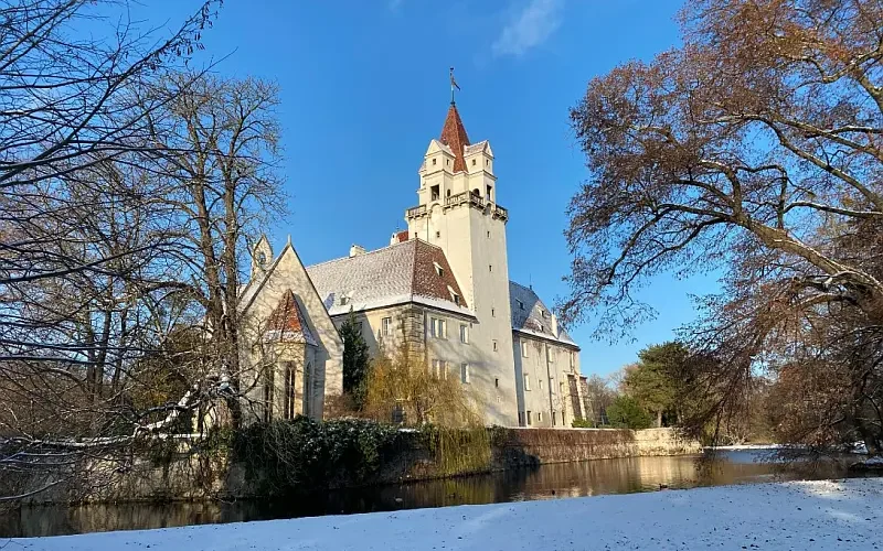 Verschneites Schloss mit Burggraben und Bäumen, im Hintergrund strahlend blauer Himmel